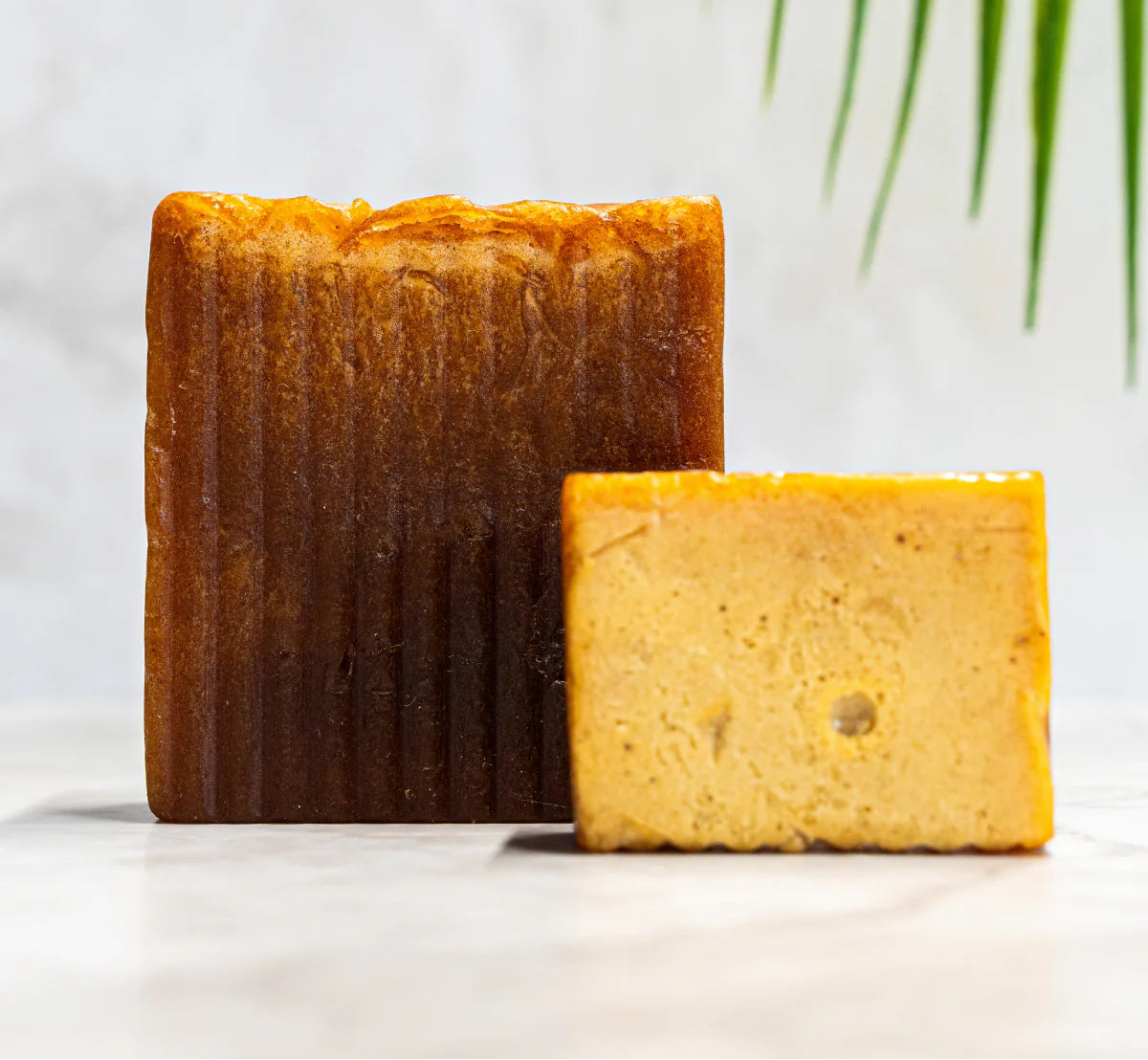 Two rectangular bars of handmade soap standing upright on a light marble-like surface, with one bar in front broken to show the textured interior. The soap has a warm amber-brown outer surface and a pale golden-yellow interior.